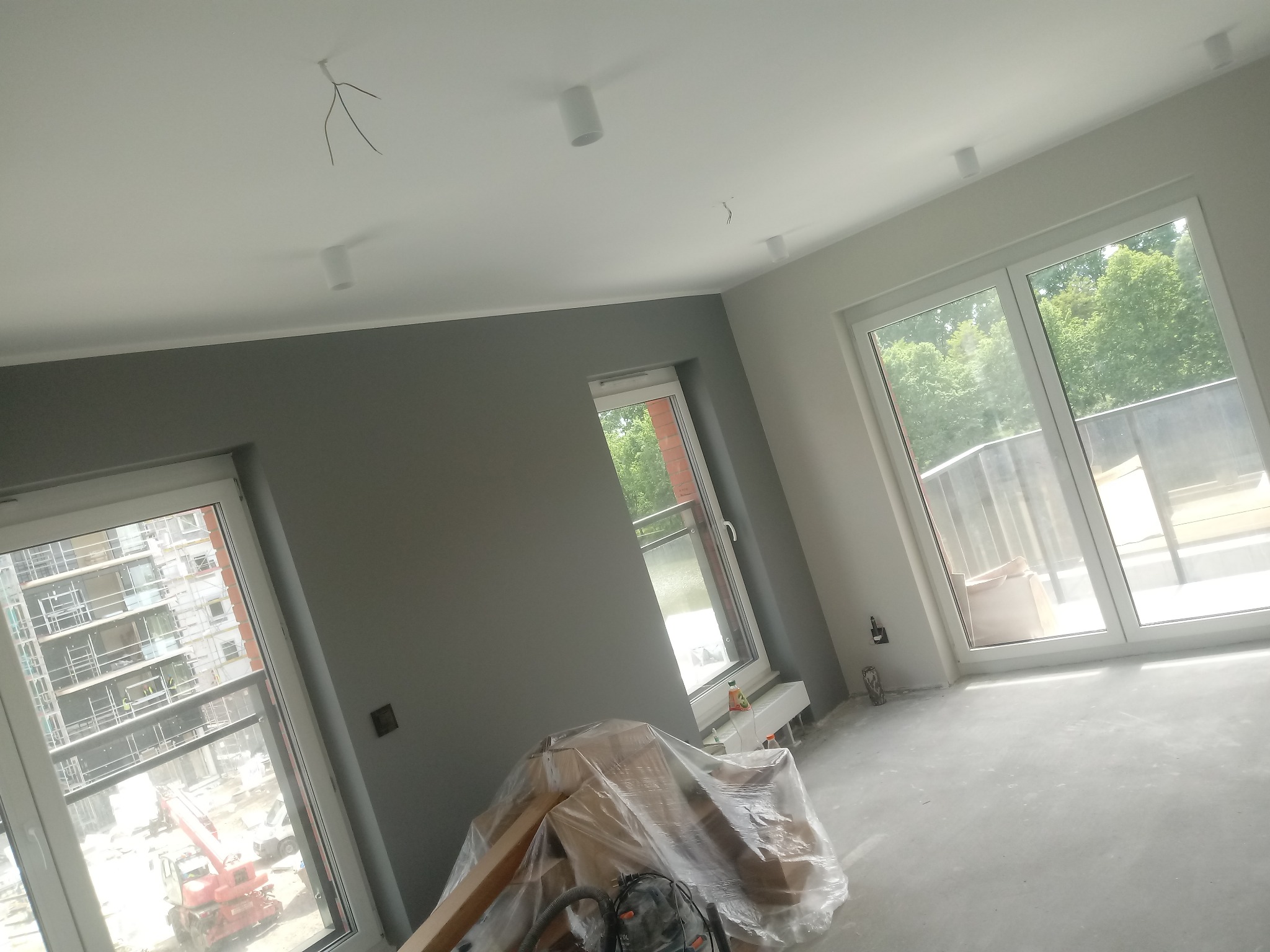 Wnętrze w trakcie remontu: szare ściany, biały sufit z zamontowanymi lampami punktowymi, okna z widokiem na zewnątrz, podłoga przygotowana do dalszych prac, materiały budowlane przykryte folią...