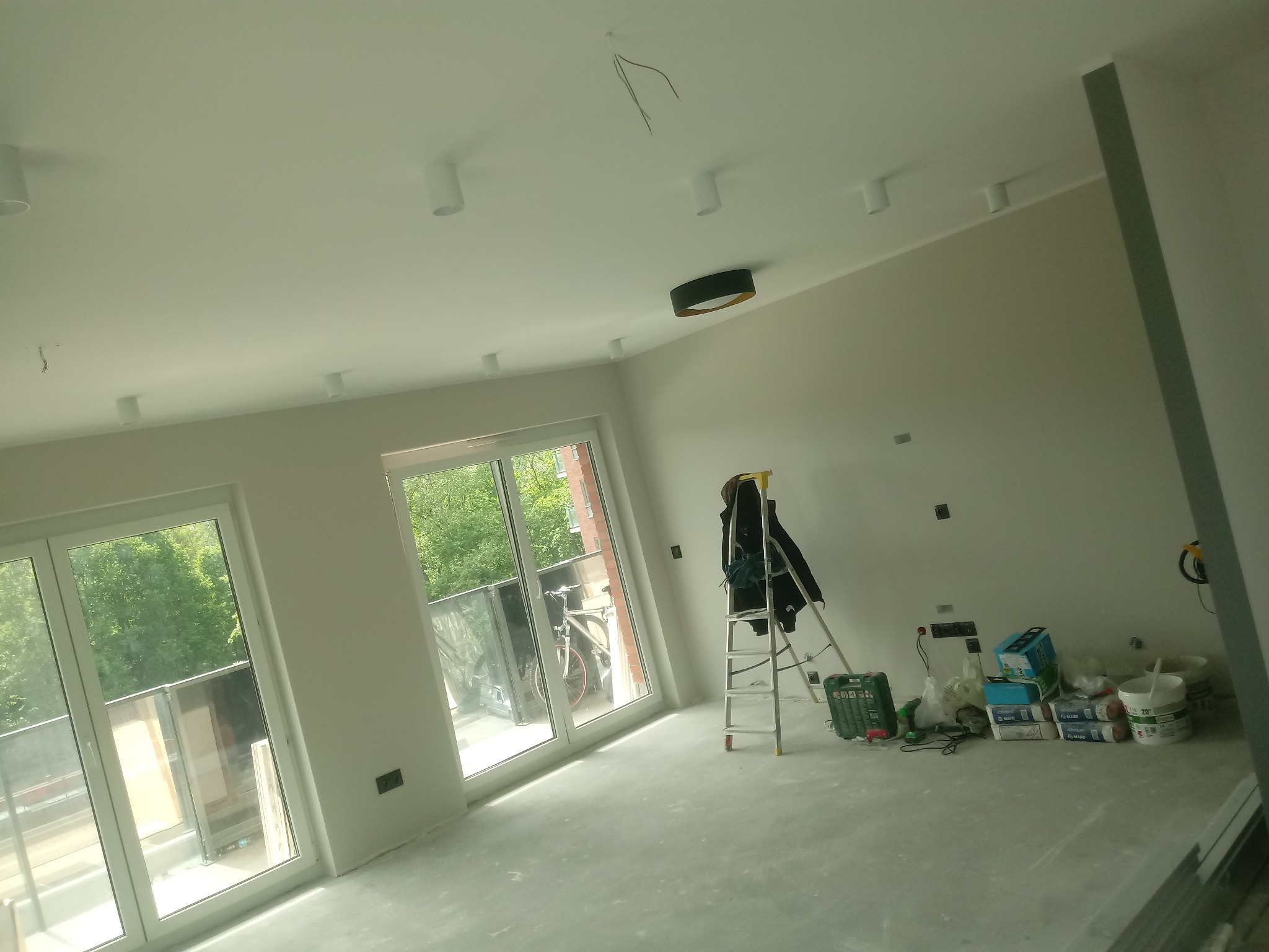 Wnętrze w trakcie remontu: świeżo pomalowane ściany, sufit z zamontowanymi lampami, drabina z kurtką, worki z zaprawą i narzędzia na podłodze przy oknie balkonowym.