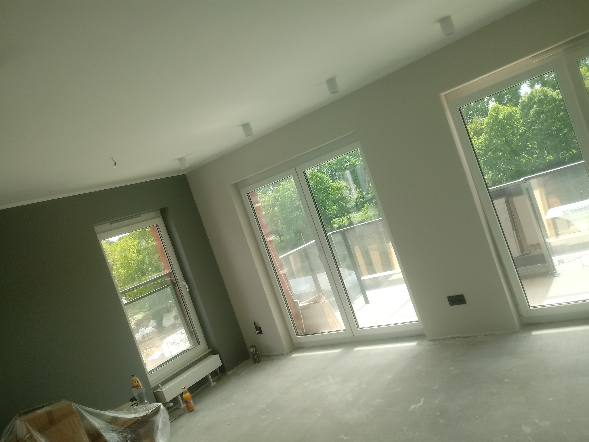 Wnętrze pokoju w trakcie remontu, widoczne trzy okna balkonowe, ściany częściowo pomalowane na szaro, podłoga przygotowana do dalszych prac wykończeniowych, widoczne butelki z napojami i zakryte...