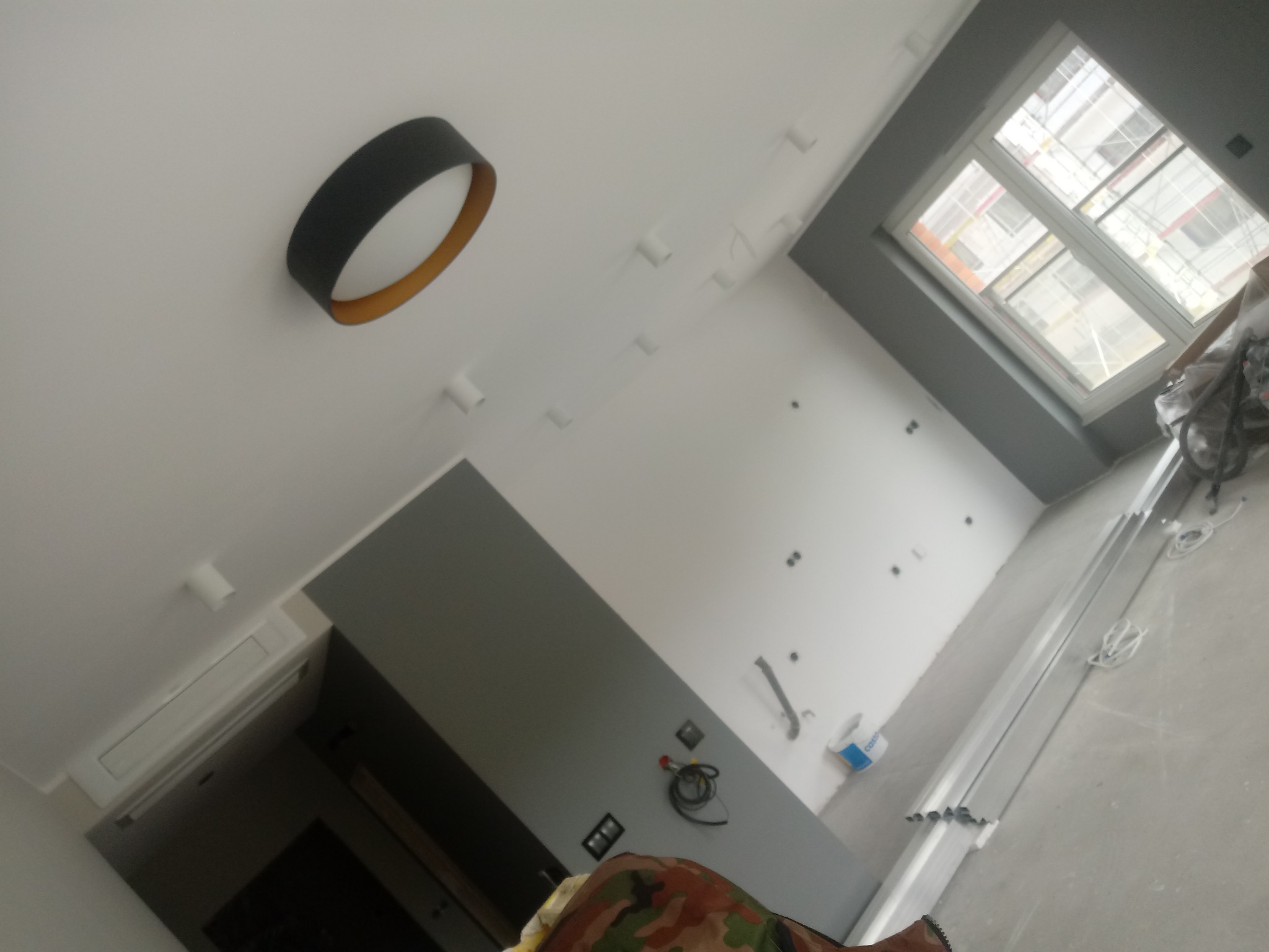 Wnętrze w trakcie prac wykończeniowych z widocznym oknem, elementami instalacji elektrycznej, zamontowanym oświetleniem sufitowym oraz klimatyzatorem, z widocznymi materiałami budowlanymi na podłodze.