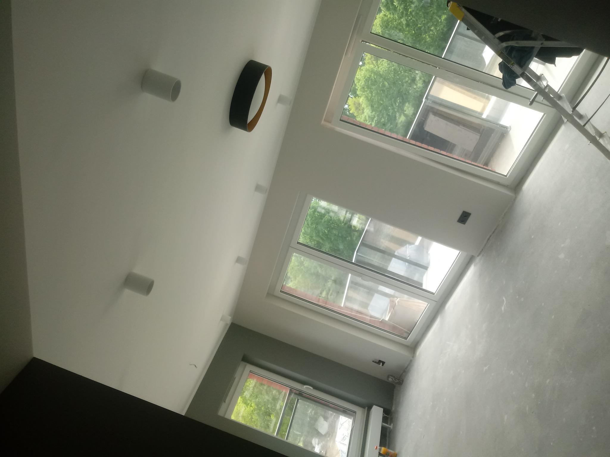 Pomieszczenie w trakcie remontu: sufit z oświetleniem punktowym i okrągłą lampą, ściany częściowo szare, częściowo przygotowane do malowania, drabina oparta o okno dachowe.