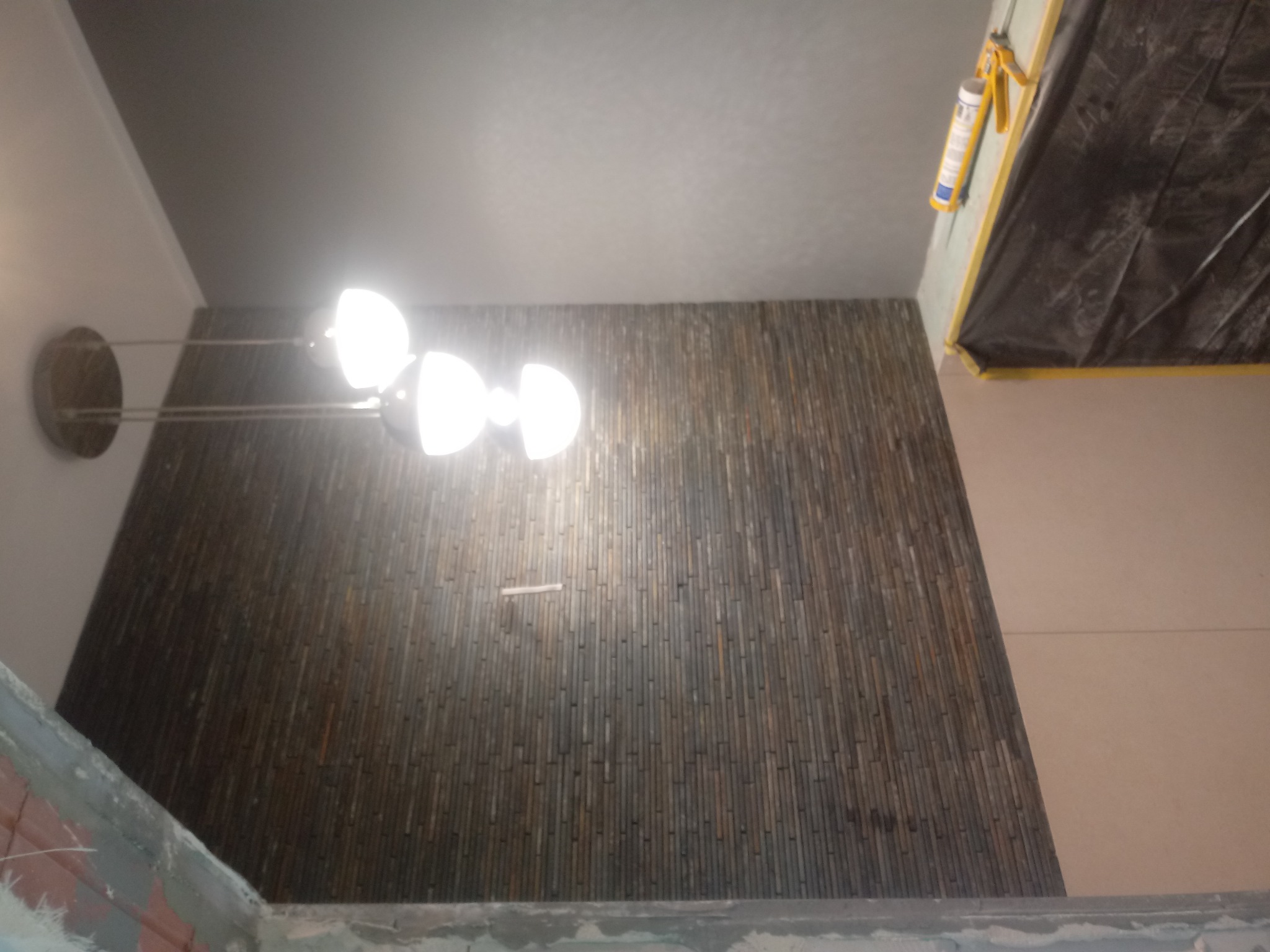 Ściana wykończona dekoracyjnymi panelami imitującymi drewno, oświetlona lampą sufitową z trzema kulistymi kloszami, widoczny fragment sufitu i zabezpieczona folią część pomieszczenia.