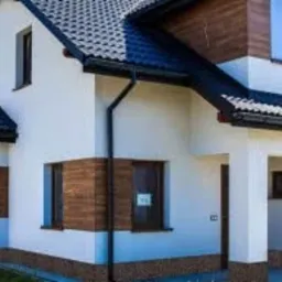 Dwukondygnacyjny dom jednorodzinny z białymi ścianami, fragmentami elewacji wykończonymi drewnem, ciemną dachówką i brązowymi oknami, widoczny z perspektywy ogrodu.