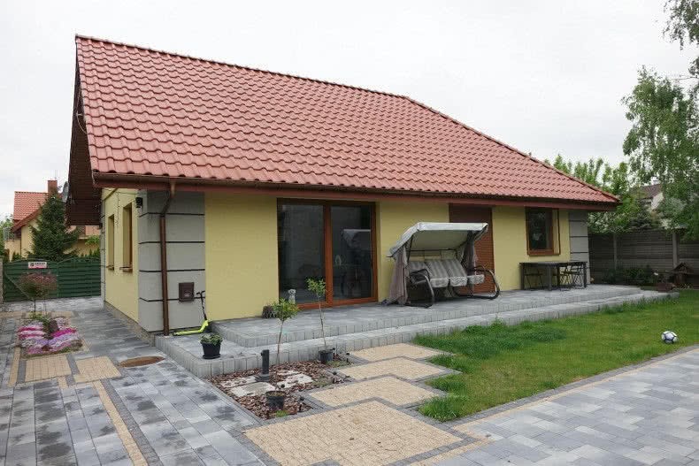 Żółty dom jednorodzinny z czerwoną dachówką, szarą elewacją na narożnikach, widoczny taras z huśtawką i kostka brukowa na podjeździe