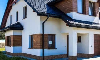 Dwukondygnacyjny dom jednorodzinny z białymi ścianami, fragmentami elewacji wykończonymi drewnem, ciemną dachówką i brązowymi oknami, widoczny z perspektywy ogrodu.