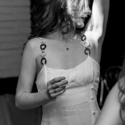 Portret kobiety w białej sukience z ozdobnymi ramiączkami, z podniesioną ręką, częściowo zasłaniającą twarz i włosy, w czarno-białej tonacji.