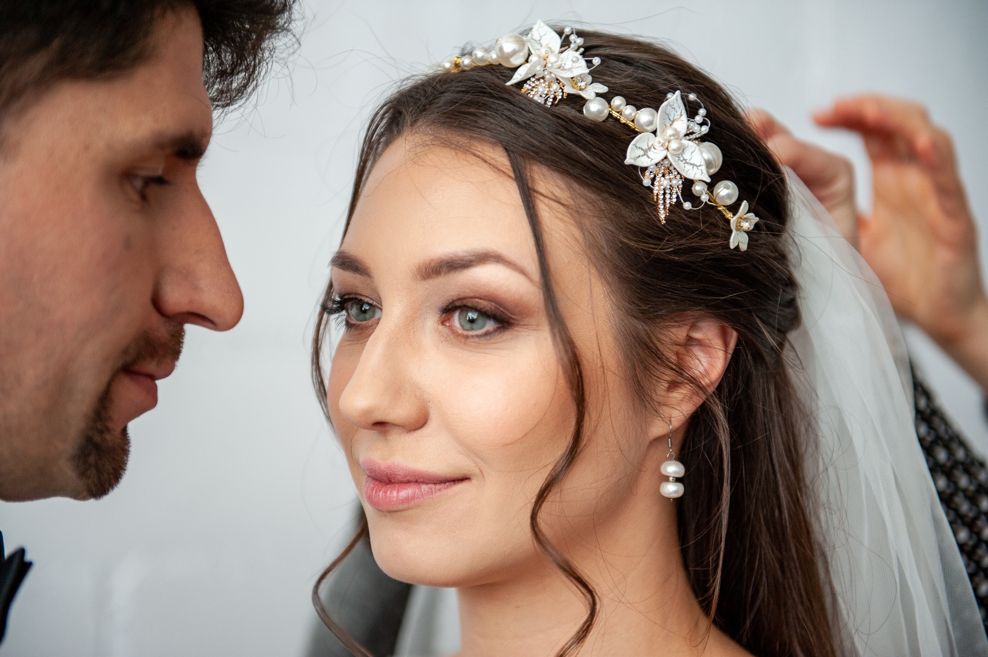 Panna młoda z delikatnym makijażem i ozdobną biżuterią we włosach, tuż przed ceremonią, wpatruje się w przyszłego męża podczas zakładania welonu.