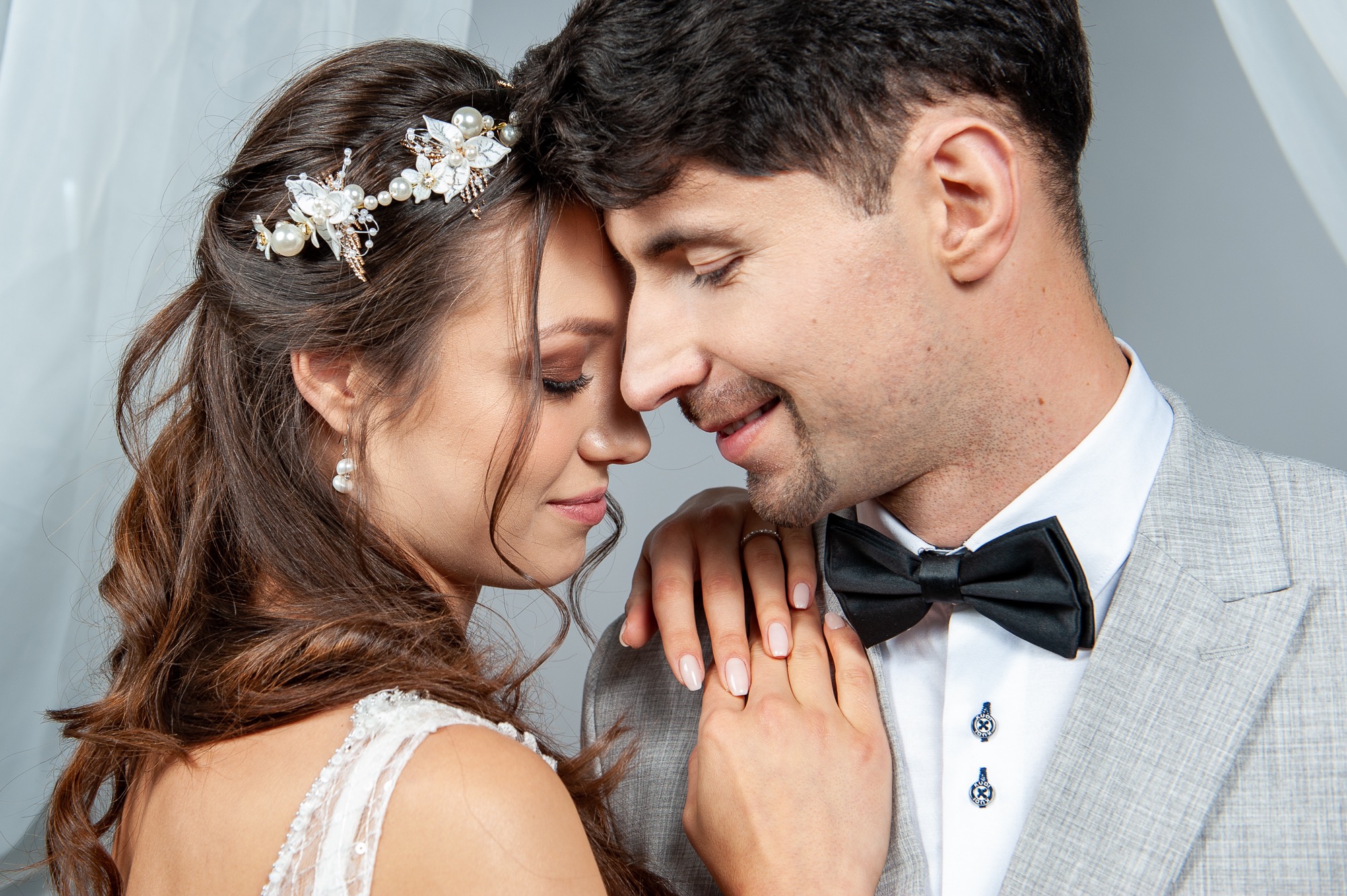 Para młoda, zbliżenie twarzy, panna młoda z ozdobą we włosach z perłami i kwiatami, pan młody w szarym garniturze i czarnej muszce, trzymają się za ręce