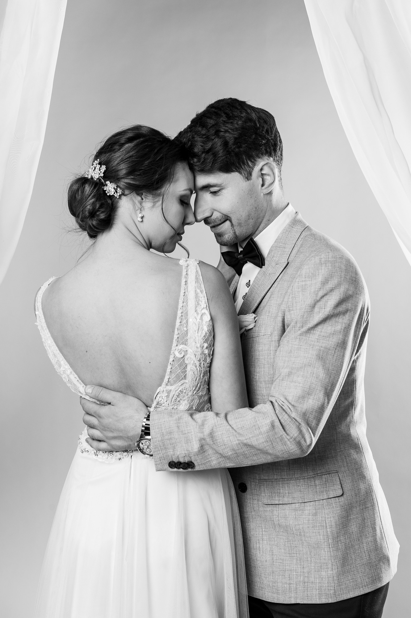 Czarno-białe zdjęcie pary młodej: panna młoda w sukni z koronkowym tyłem i upięciem z ozdobami, pan młody w szarej marynarce i muszce, obejmujący się czule na neutralnym tle z fragmentem białej...