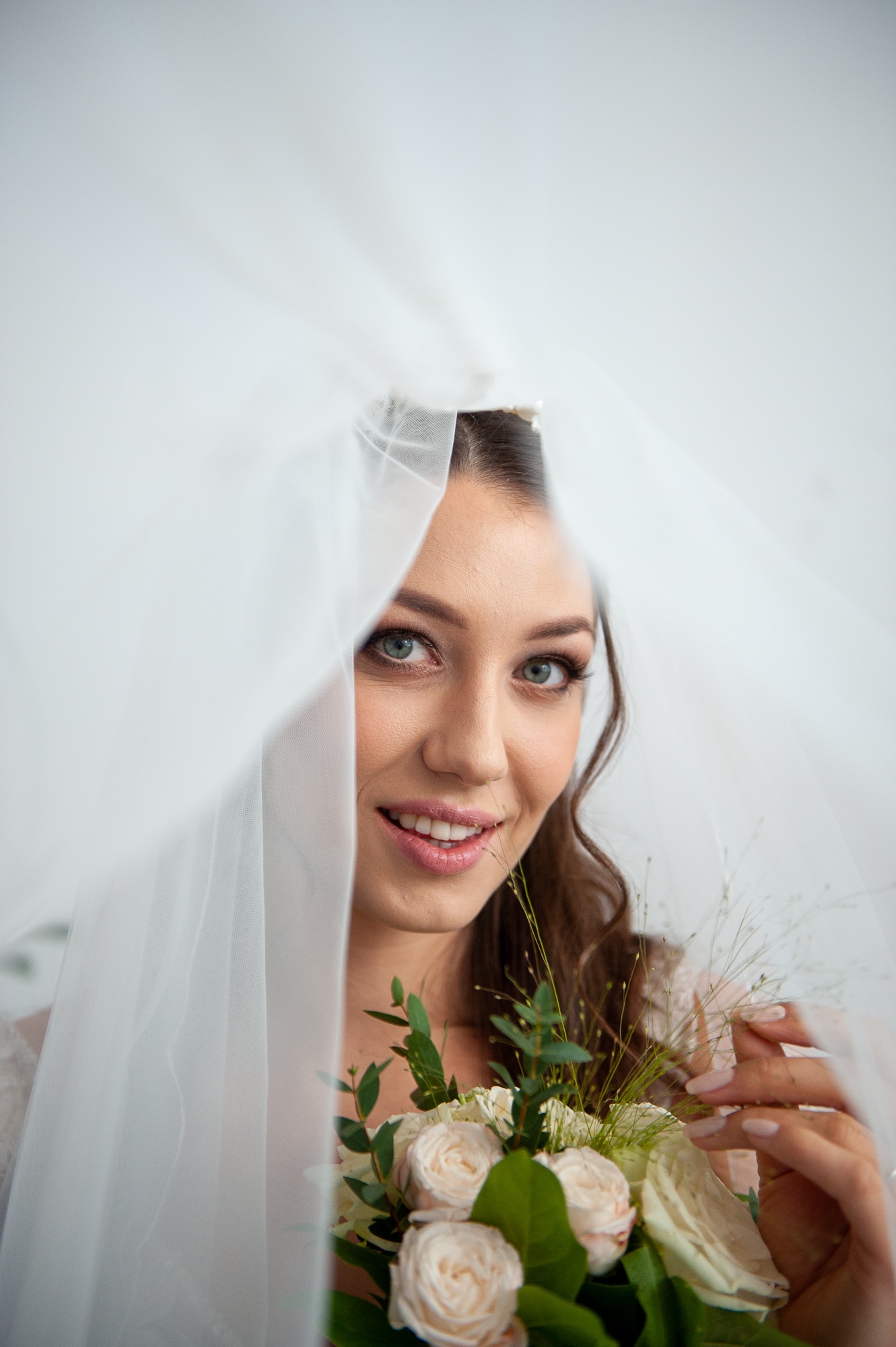 Uśmiechnięta panna młoda z delikatnym makijażem, spoglądająca spod białego welonu, trzymająca bukiet z białych róż i zielonych liści.
