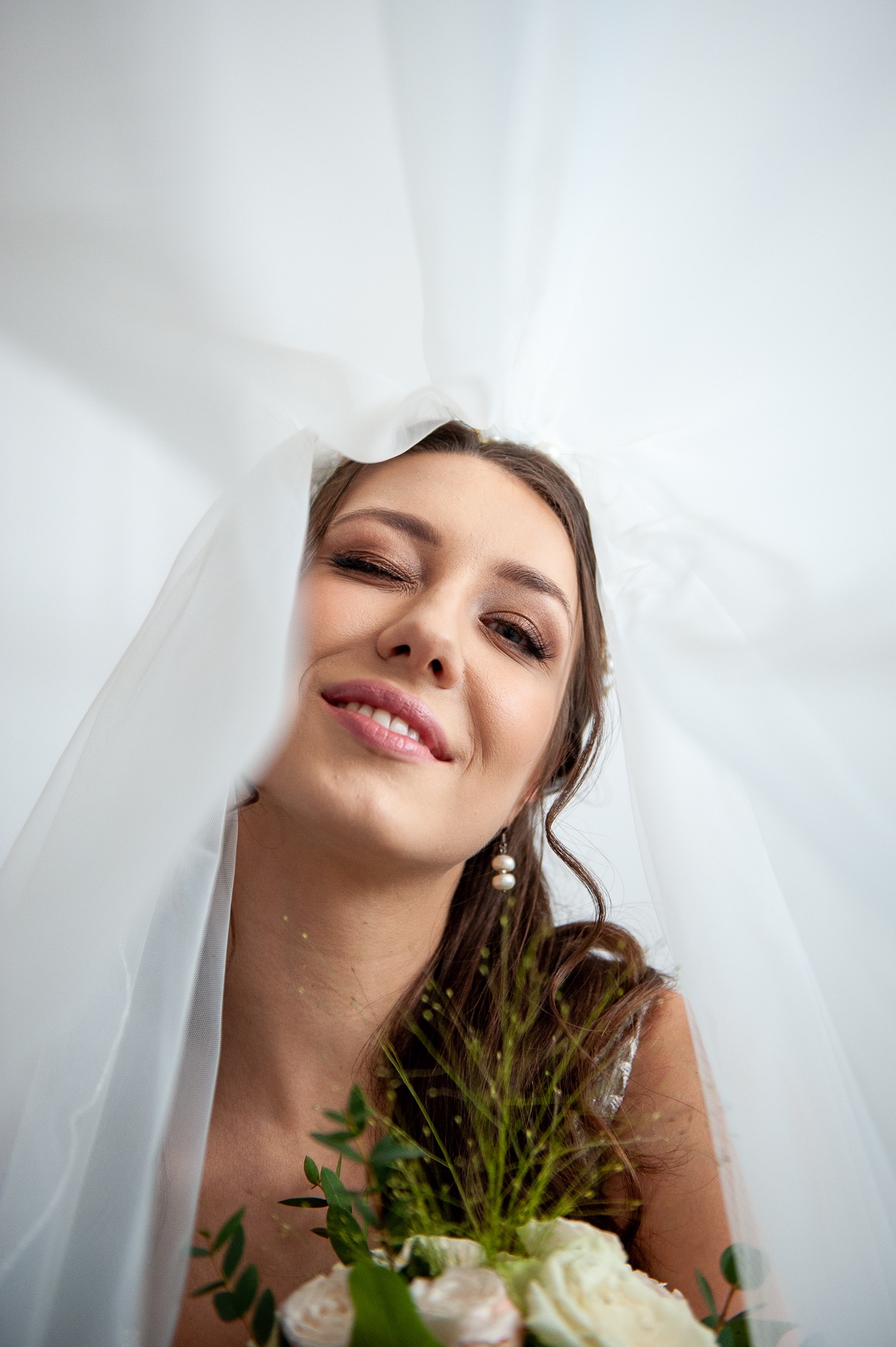 Uśmiechnięta panna młoda w białym welonie, z zalotnie przymrużonym okiem i bukietem białych róż, portret z dołu.