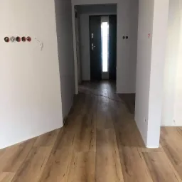 Korytarz w trakcie prac wykończeniowych: położone panele podłogowe o wzorze drewna, surowe ściany z widocznymi puszkami elektrycznymi i przewodami, czarne drzwi wejściowe z pionowym przeszkleniem.