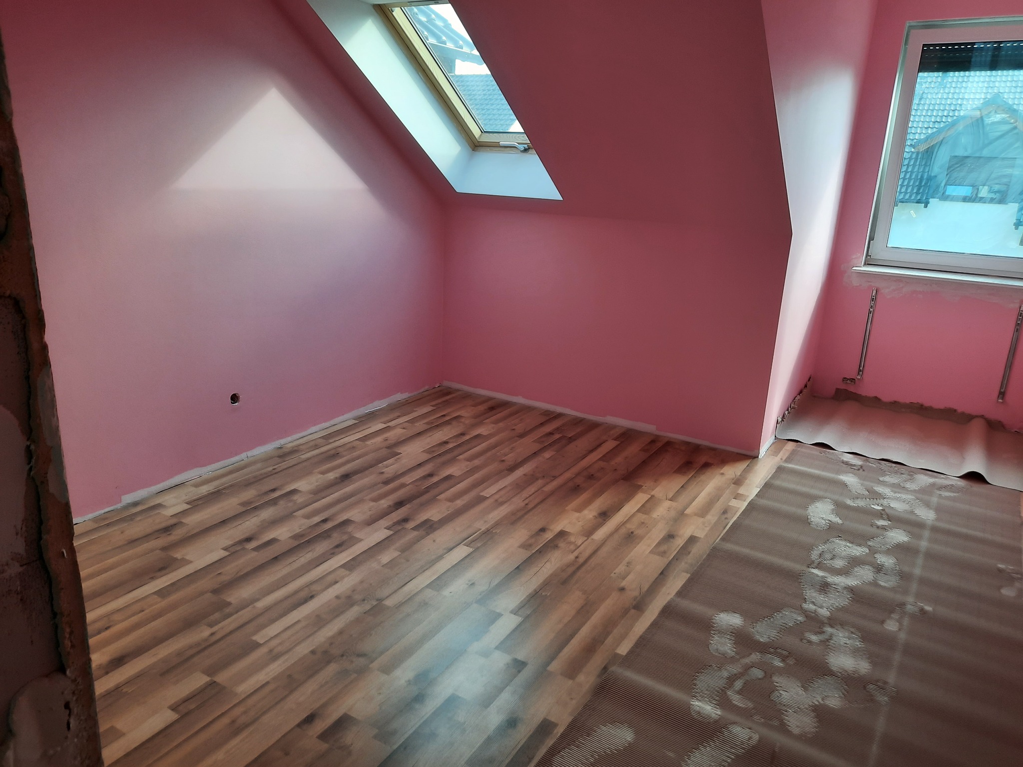 Pokój na poddaszu z oknem dachowym i nową podłogą z paneli, ściany pomalowane na różowo, widoczne ślady po butach na folii ochronnej.