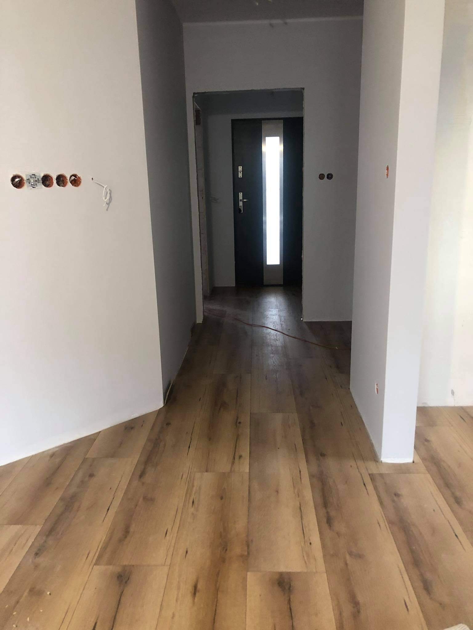 Korytarz w trakcie prac wykończeniowych: położone panele podłogowe o wzorze drewna, surowe ściany z widocznymi puszkami elektrycznymi i przewodami, czarne drzwi wejściowe z pionowym przeszkleniem.