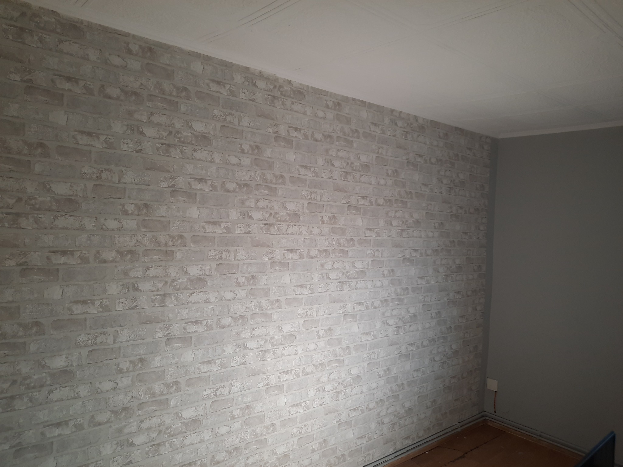 Ściana wykończona tapetą imitującą białą cegłę, widoczny fragment sufitu z listwą oraz szara ściana w rogu pokoju.