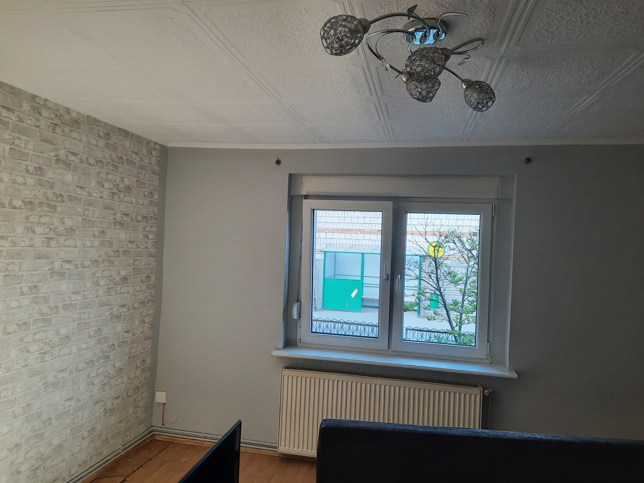 Pokój po odświeżeniu: ściana z dekoracyjną cegłą w odcieniach szarości, białe okno z widokiem na zewnątrz, grzejnik pod oknem, designerska lampa sufitowa.