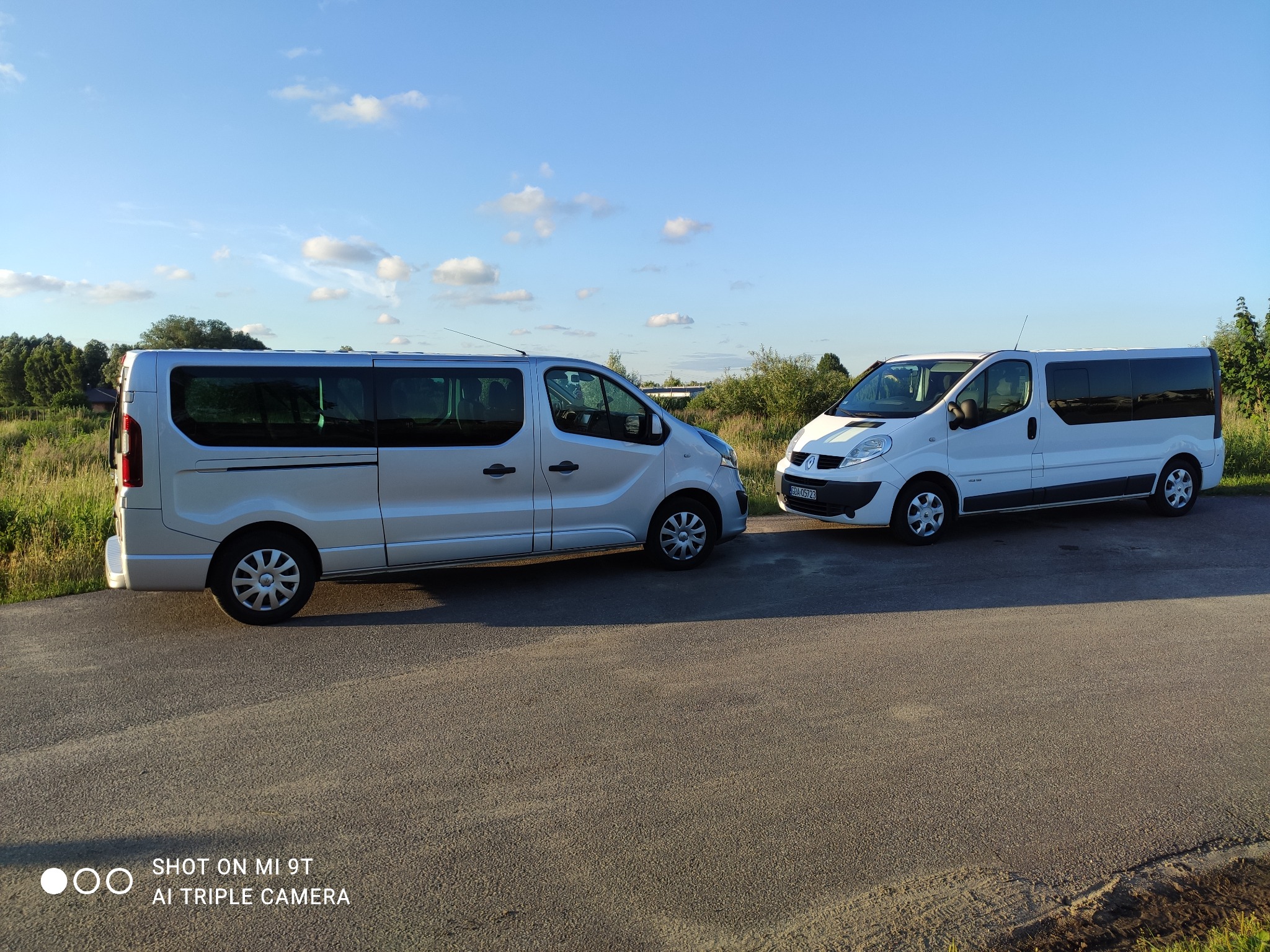 Dwa busy: srebrny i biały, stojące na asfalcie, z widocznym zielonym otoczeniem i błękitnym niebem z chmurami w tle, sugerujące gotowość do przewozu osób.
