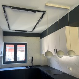 Dan Elektro Instal - Nowoczesna kuchnia z czarnymi blatami, białymi szafkami i oryginalnym oświetleniem sufitowym w kształcie ramy. Widoczne trzy wiszące lampy kuliste.