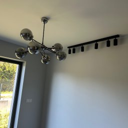 Dan Elektro Instal - Nowoczesne oświetlenie sufitowe: chromowany żyrandol kulisty i czarna listwa z reflektorami, widoczne na tle białego sufitu i ściany, z oknem w tle.