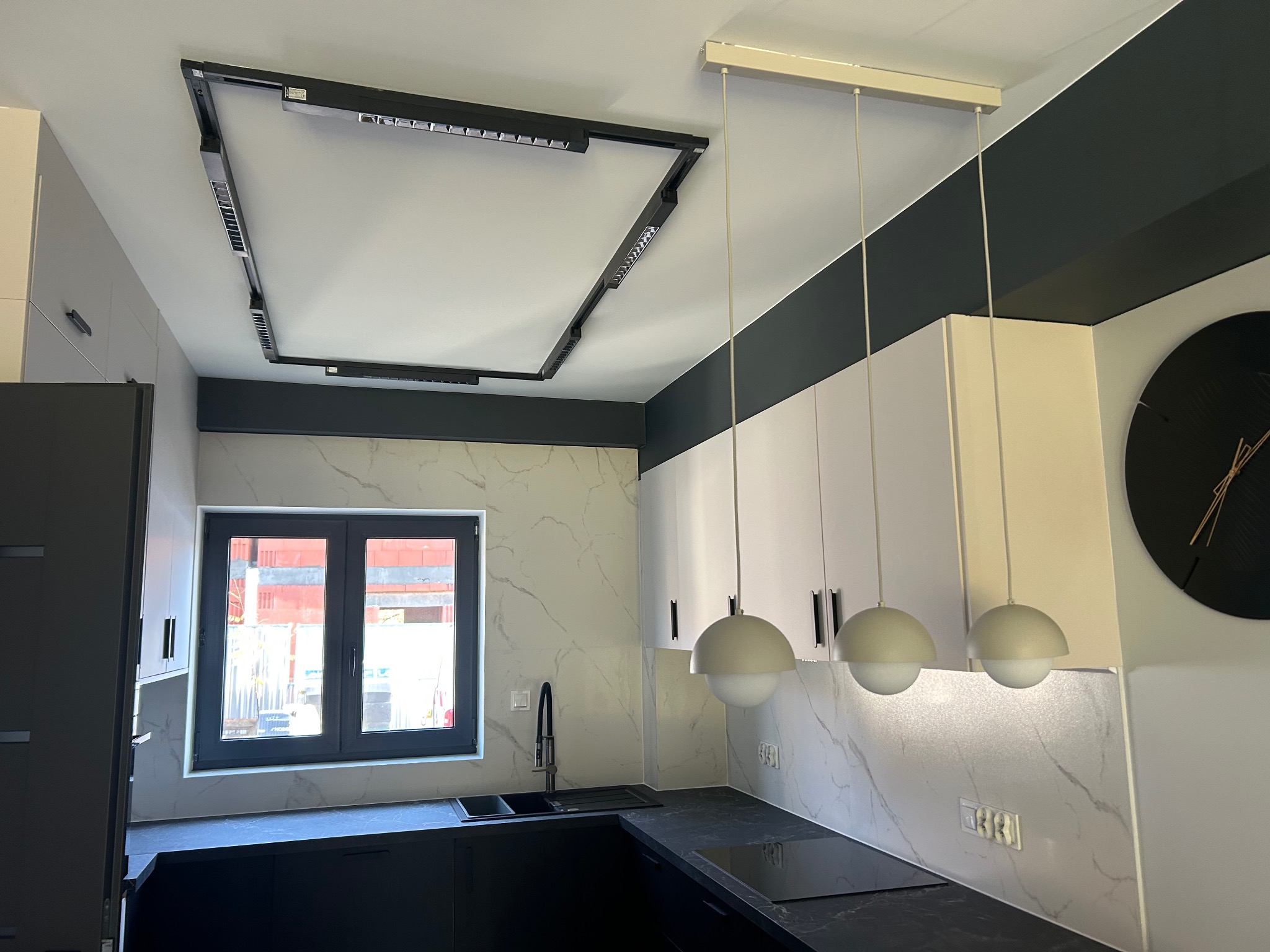 Nowoczesna kuchnia z czarnymi blatami, białymi szafkami i oryginalnym oświetleniem sufitowym w kształcie ramy. Widoczne trzy wiszące lampy kuliste.