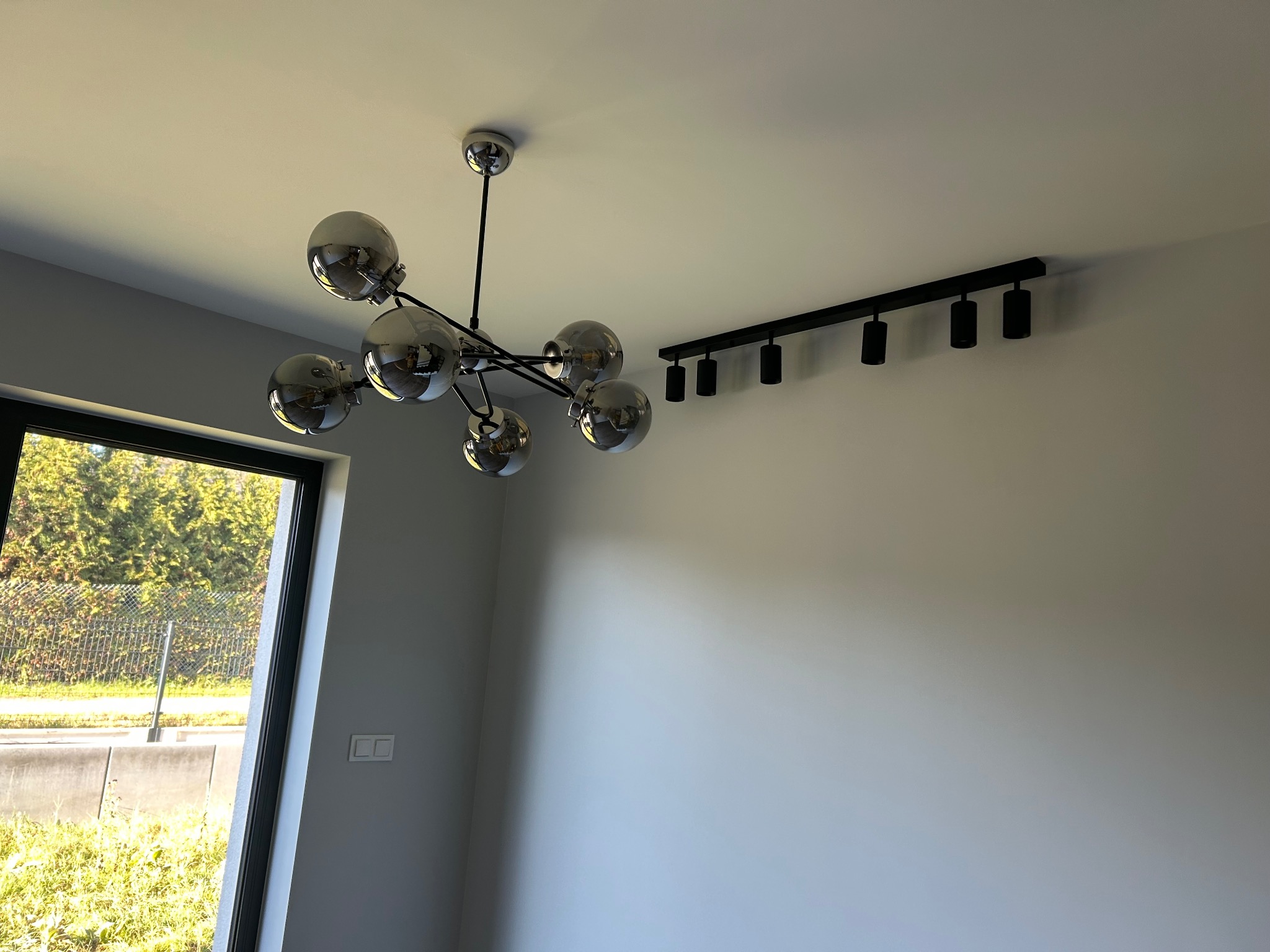 Nowoczesne oświetlenie sufitowe: chromowany żyrandol kulisty i czarna listwa z reflektorami, widoczne na tle białego sufitu i ściany, z oknem w tle.