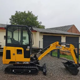 Żółto-czarna mini koparka JCB 16C-1, stojąca na żwirowym podłożu, z widocznymi elementami konstrukcyjnymi i łyżką, na tle magazynu z szarymi drzwiami.