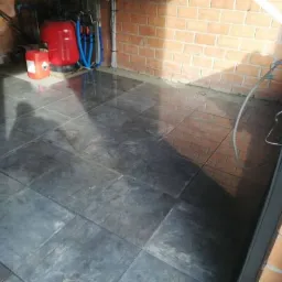 Świeżo położona, ciemnoszara podłoga z płytek w pomieszczeniu z ceglanymi ścianami, widoczny zbiornik ciśnieniowy i kanister z etykietą ostrzegawczą.