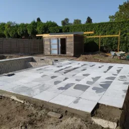 Budowa basenu ogrodowego z szarych płyt betonowych, widoczny drewniany domek narzędziowy i fragment ogrodu.