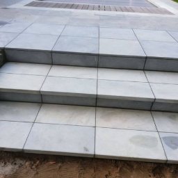 Marcin Sarnowski - Trzy stopnie schodów wyłożone jasnoszarymi, kwadratowymi płytkami, widoczne niedokończone obrzeża z kamienia i ziemi.