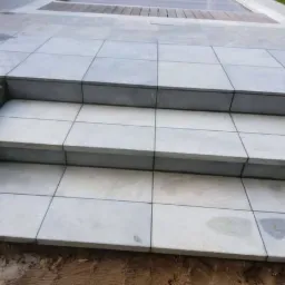 Trzy stopnie schodów wyłożone jasnoszarymi, kwadratowymi płytkami, widoczne niedokończone obrzeża z kamienia i ziemi.