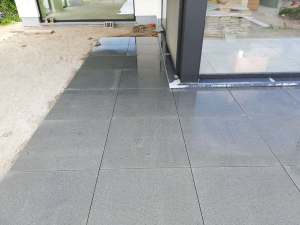 Ułożona, ciemnoszara posadzka z płytek na zewnątrz budynku, widoczne okno tarasowe i piasek obok.