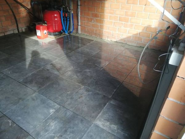 Świeżo położona, ciemnoszara podłoga z płytek w pomieszczeniu z ceglanymi ścianami, widoczny zbiornik ciśnieniowy i kanister z etykietą ostrzegawczą.