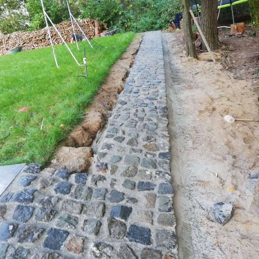 Układanie ścieżki z kostki brukowej, widoczny fragment ułożonej nawierzchni, przygotowane podłoże z piasku i obrzeża, trawnik z boku, narzędzie do ubijania.