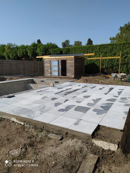Budowa basenu ogrodowego z szarych płyt betonowych, widoczny drewniany domek narzędziowy i fragment ogrodu.