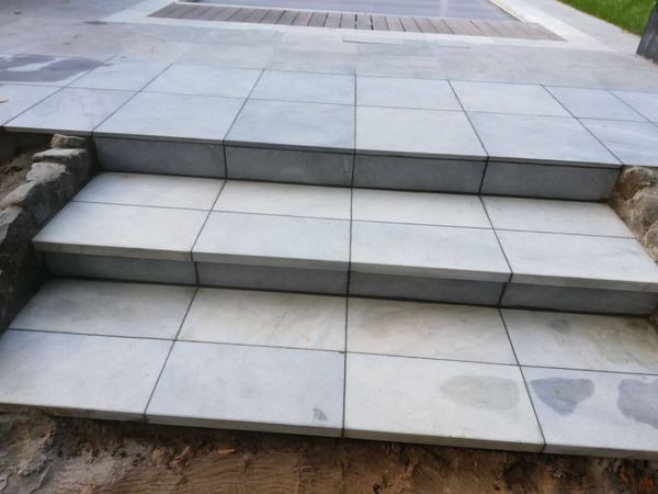 Trzy stopnie schodów wyłożone jasnoszarymi, kwadratowymi płytkami, widoczne niedokończone obrzeża z kamienia i ziemi.