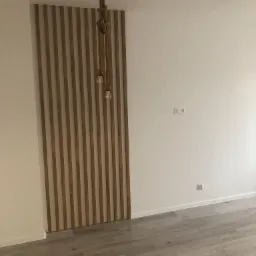 Panel dekoracyjny z drewnianych listew na ścianie, lampa wisząca z grubego sznura i żarówkami Edisona, szare panele podłogowe, białe ściany z gniazdkami.