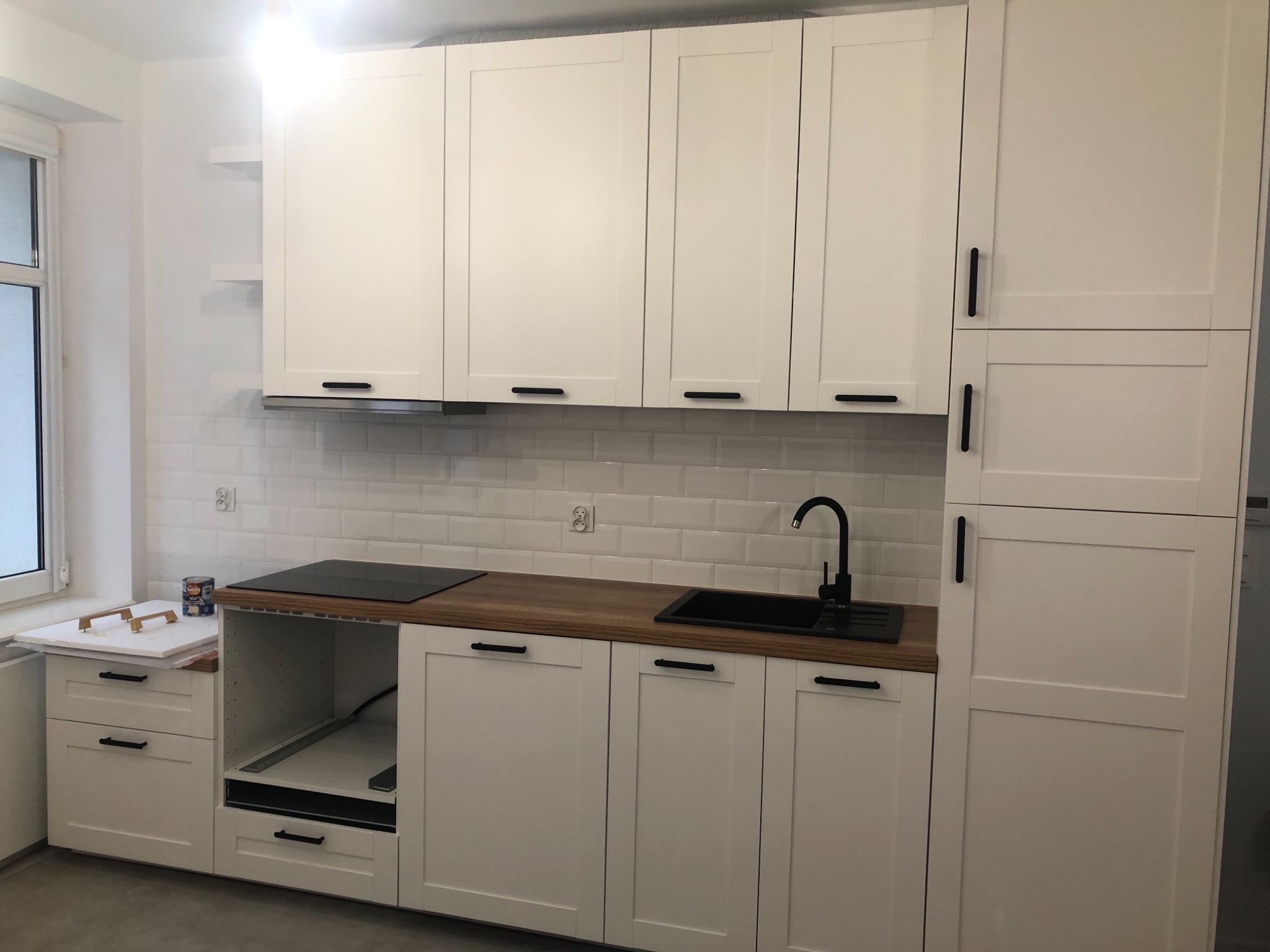 Nowoczesna, jasna kuchnia z białymi szafkami z czarnymi uchwytami, drewnianym blatem i białymi płytkami na ścianie, widoczne przygotowania do montażu dodatkowych elementów.