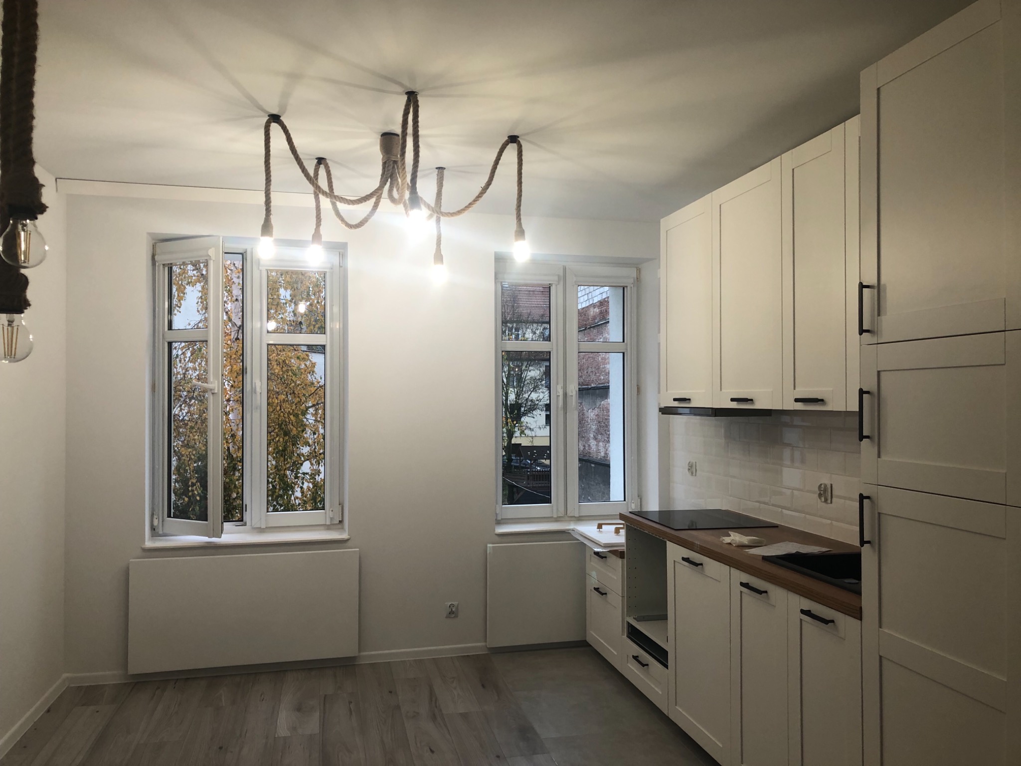 Jasna kuchnia po remoncie z białymi szafkami, drewnianym blatem i oryginalną lampą z liny jutowej zwisającą z sufitu.