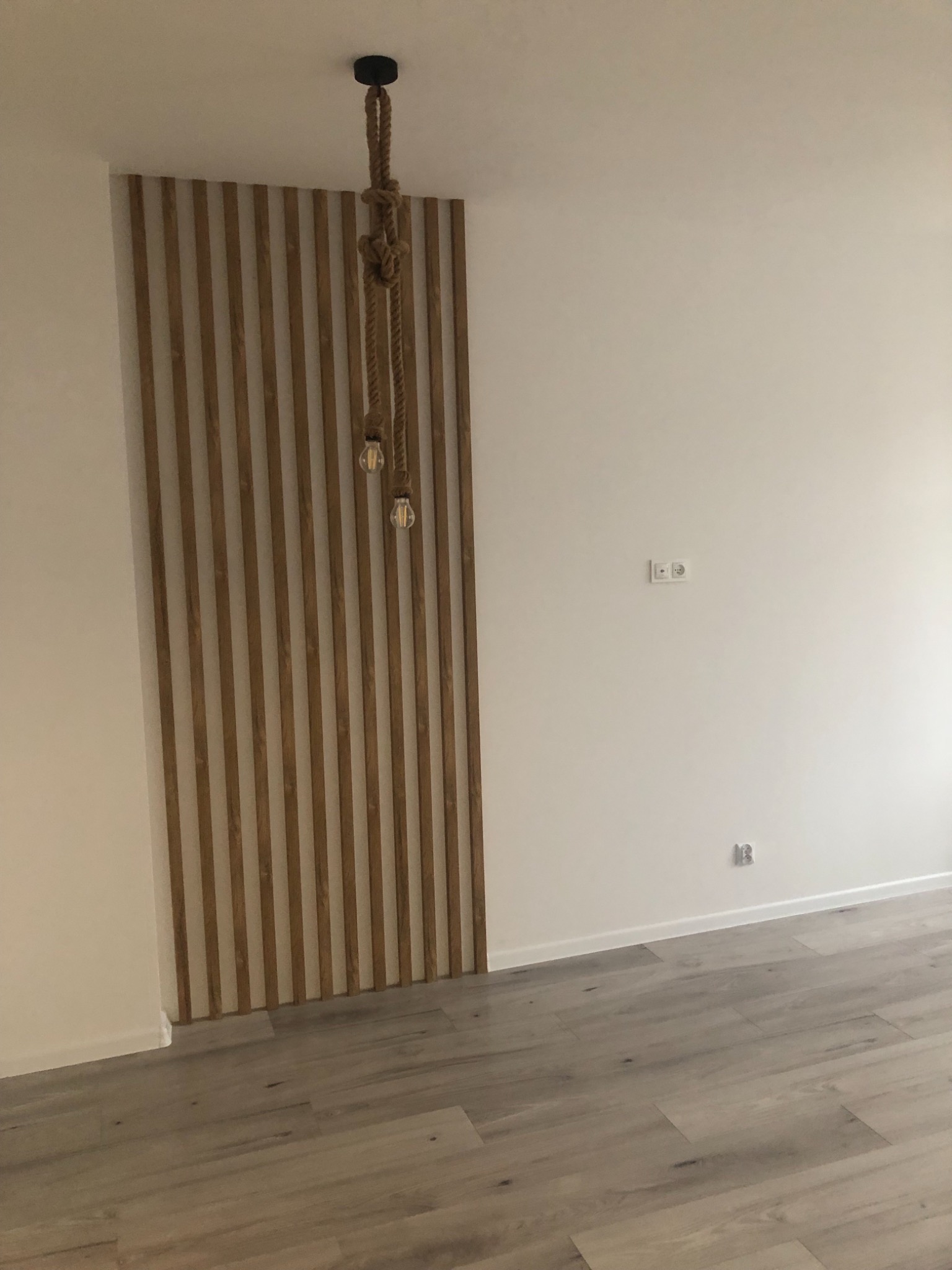 Panel dekoracyjny z drewnianych listew na ścianie, lampa wisząca z grubego sznura i żarówkami Edisona, szare panele podłogowe, białe ściany z gniazdkami.