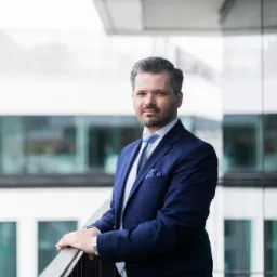 Portret mężczyzny w granatowym garniturze na balkonie nowoczesnego biurowca z przeszkloną fasadą.