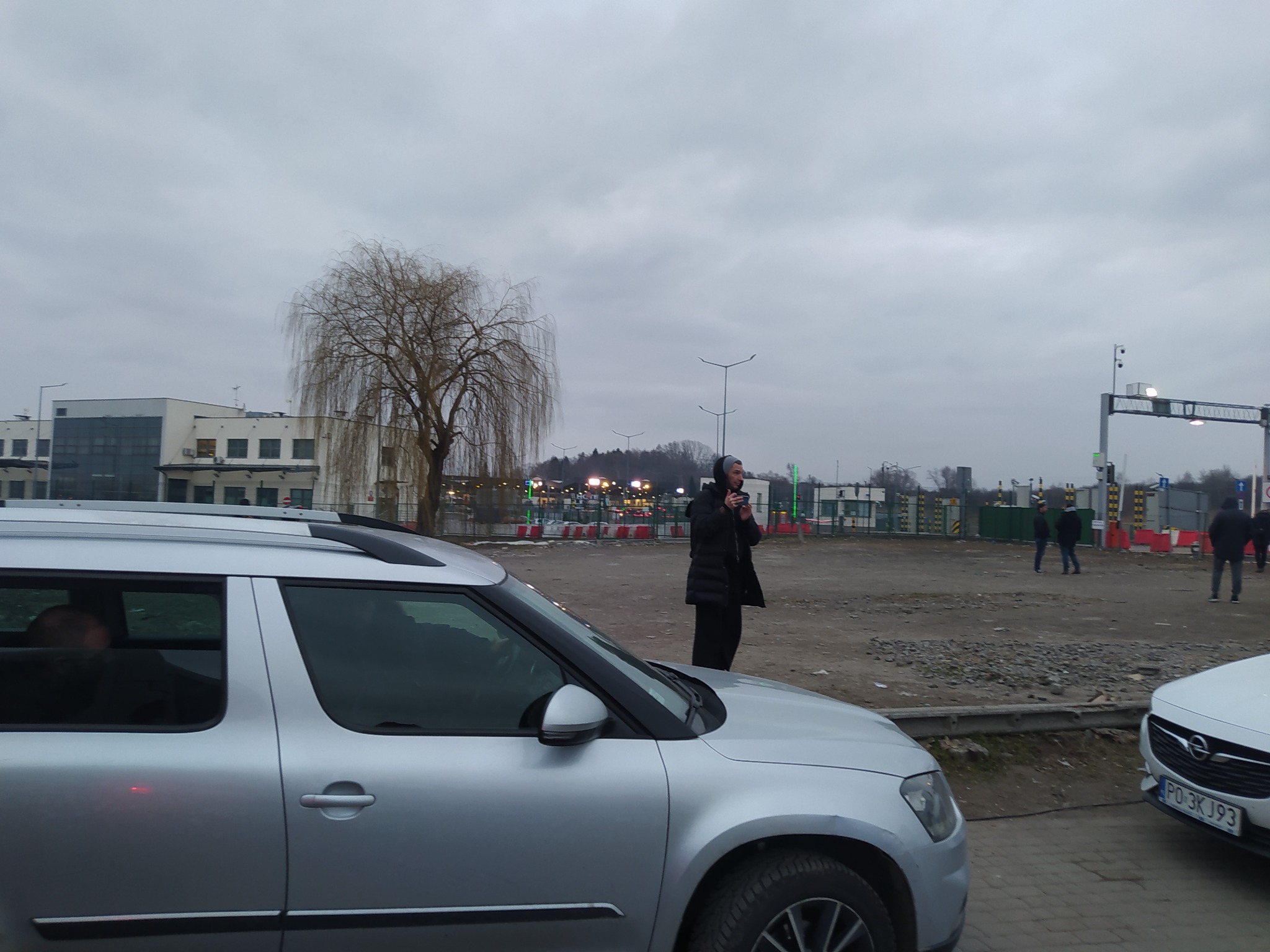 Srebrny SUV zaparkowany na szarym placu, osoba w ciemnym płaszczu robi zdjęcie telefonem, w tle budynek i szpaler świateł.