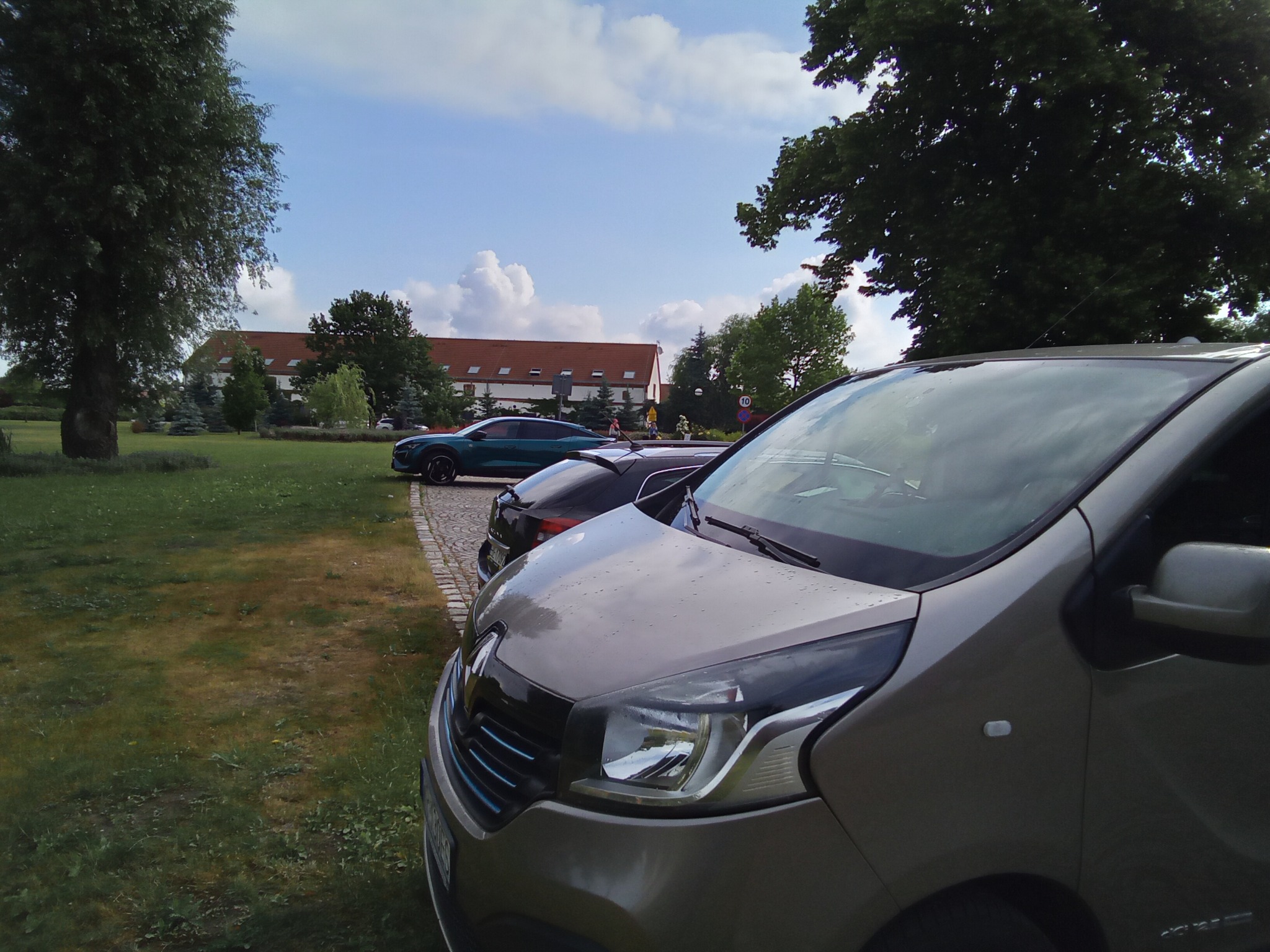 Szary van Renault z widocznymi reflektorami i zielony samochód osobowy w tle, na parkingu z kostki brukowej przed budynkiem z czerwoną dachówką i zielonym trawnikiem.