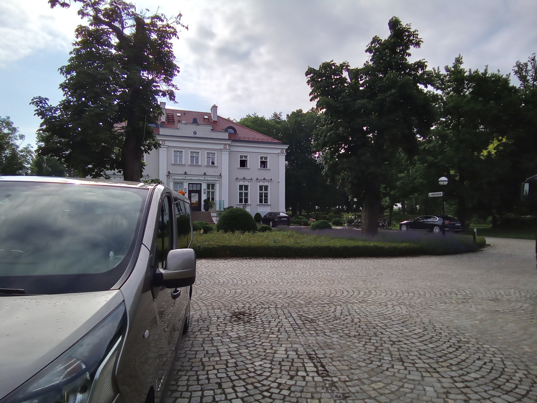 Szary van na brukowanym podjeździe przed budynkiem z kolumnami, w tle zieleń i inne zaparkowane samochody.