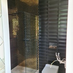 Maciej Pawłowski - Nowoczesna łazienka z kabiną prysznicową wyłożoną złotą mozaiką w kształcie plastra miodu, kontrastującą z czarnymi, podłużnymi płytkami na ścianie i drewnopodobną podłogą, z widoczną wiszącą...