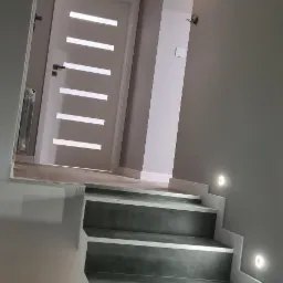 Nowoczesne schody z szarego betonu architektonicznego z podświetleniem LED przy ścianie w jasnym wnętrzu, białe drzwi z poziomymi przeszkleniami w tle.