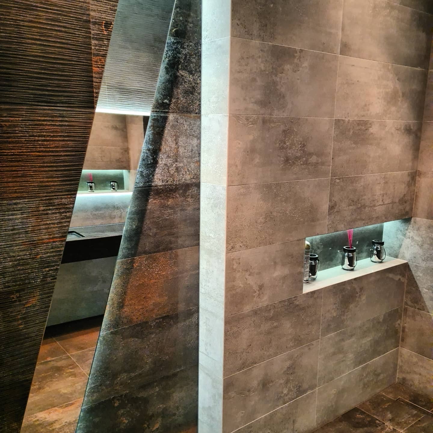 Nowoczesna łazienka z szarymi, podłużnymi płytkami, oświetloną wnęką z dekoracjami i asymetrycznym lustrem z elementami drewna.