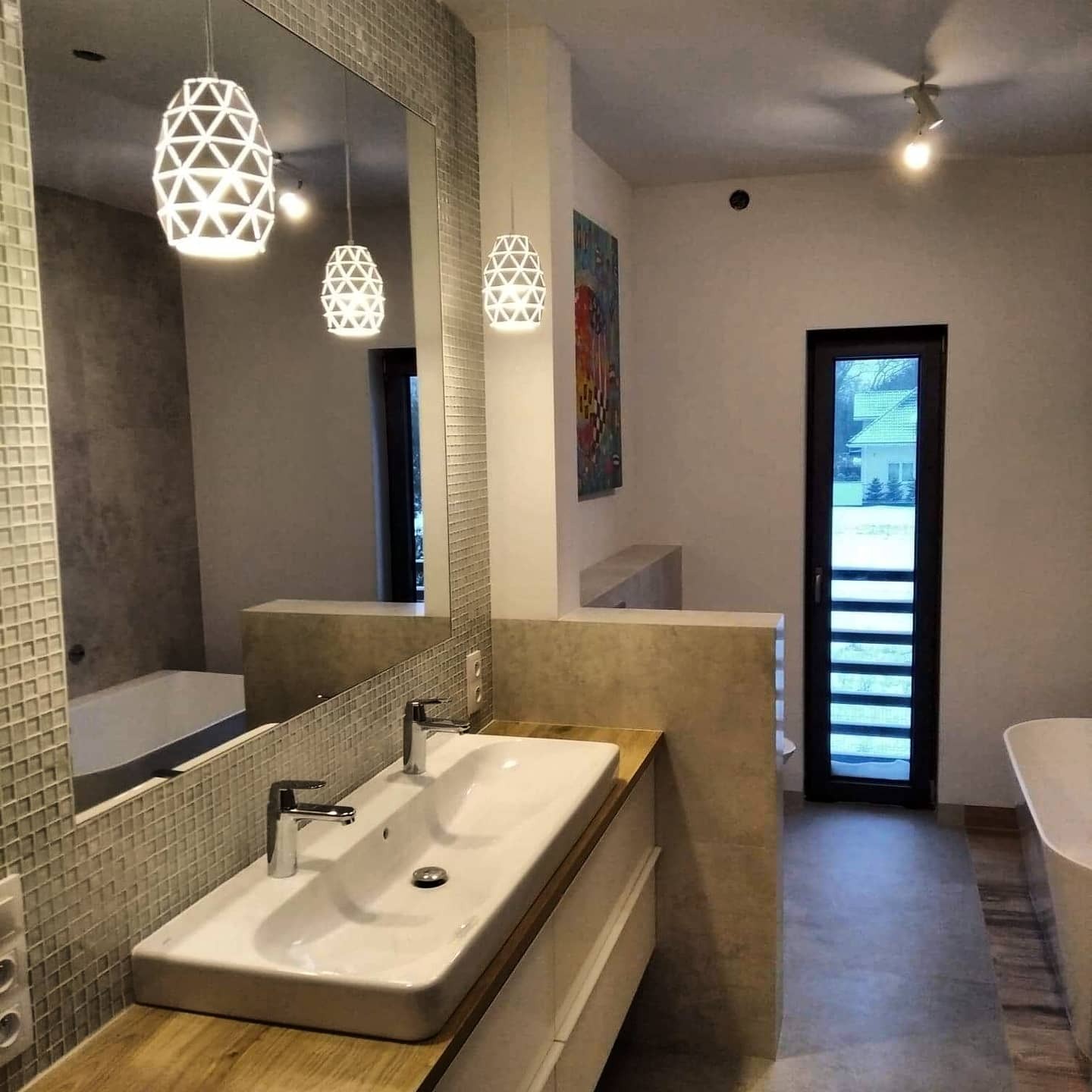 Nowoczesna łazienka z podwójną umywalką nablatową na drewnianym blacie, lustrem z mozaikową ramą, geometrycznymi lampami wiszącymi i widoczną wanną w tle.
