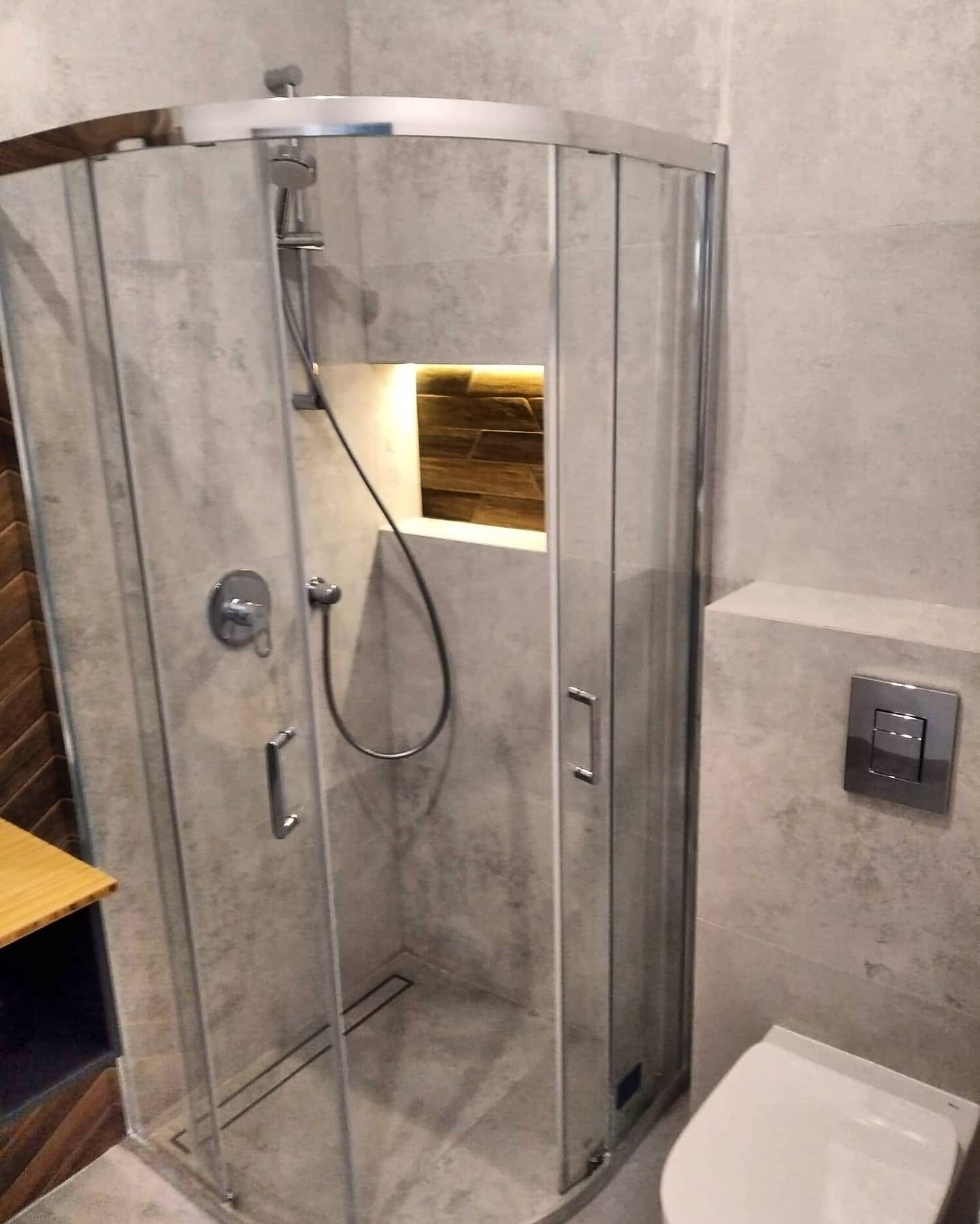 Szklana kabina prysznicowa narożna z brodzikiem liniowym, szare płytki imitujące beton, drewniana wnęka z oświetleniem, wisząca toaleta z chromowanym przyciskiem spłukiwania.