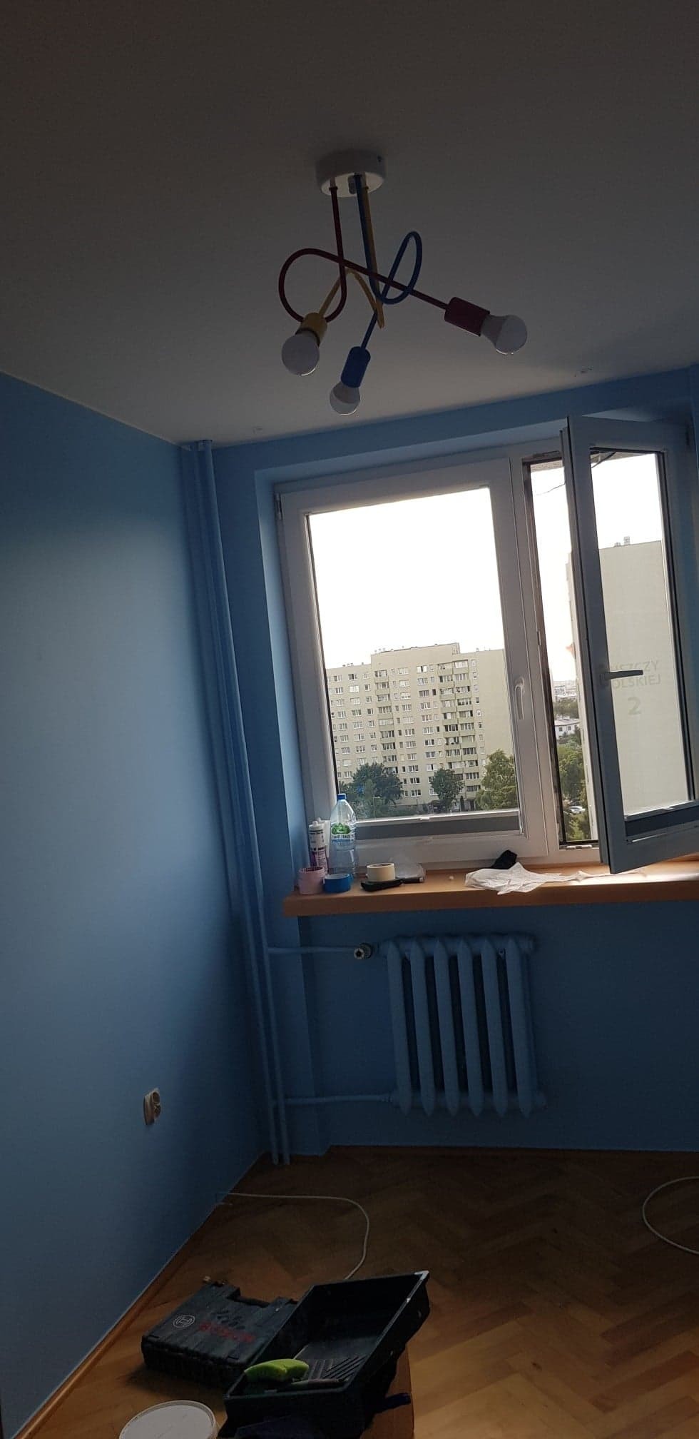 Pomalowany na niebiesko pokój z otwartym oknem, przez które widać blok, pod oknem grzejnik, na podłodze akcesoria malarskie i parkiet w jodełkę.