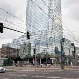 Szklany biurowiec w miejskiej scenerii z ulicą, tramwajami i przechodniami w pochmurny dzień.